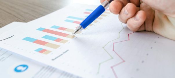 A close-up of a hand with a pen analyzing data on colorful bar and line charts on paper.